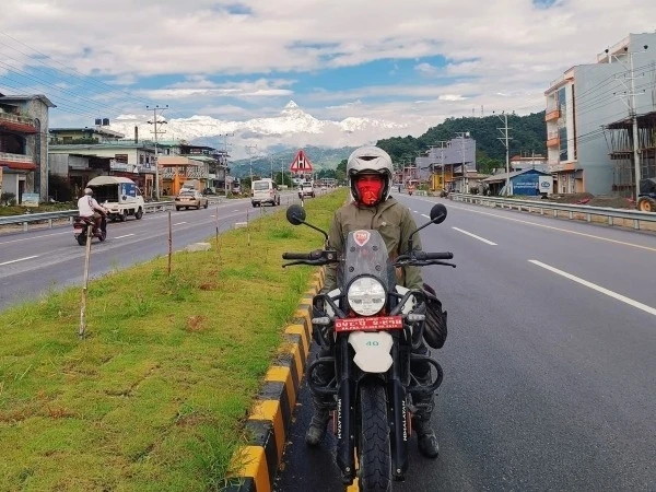 Motorbike rider on a Royal Enfield Himalayan along a highway in Nepal, with snow-capped Himalayan peaks in the background.